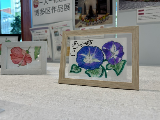 一人一花運動博多区作品展の写真。木製のフレームに入った青紫色の朝顔の花のイラストが飾られている。